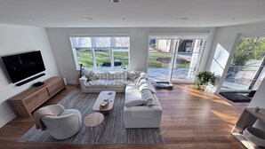 A modern living room with a sectional sofa, a flat-screen TV, a wooden cabinet, and large windows offering a view of a green landscape. - New construction, 4-bedroom house with, fitness room in lovely Bridgehampton (Bridgehampton)