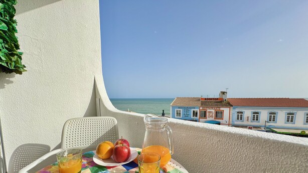 Un balcon avec une table préparée pour le petit-déjeuner, comprenant un beignet, des fruits et du jus, donnant sur une ville côtière et la mer.