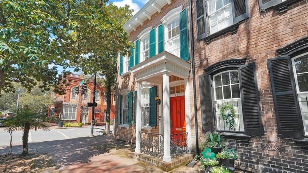A brick building with a red door and green shutters, a white porch, and a brick pathway.