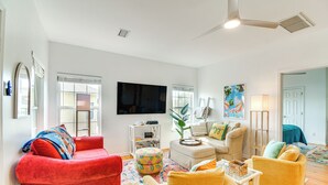 Un salón amplio con un sofá rojo, un sillón amarillo y un sofá blanco. Hay un televisor de pantalla plana montado en la pared, una mesa de centro y una lámpara de pie.