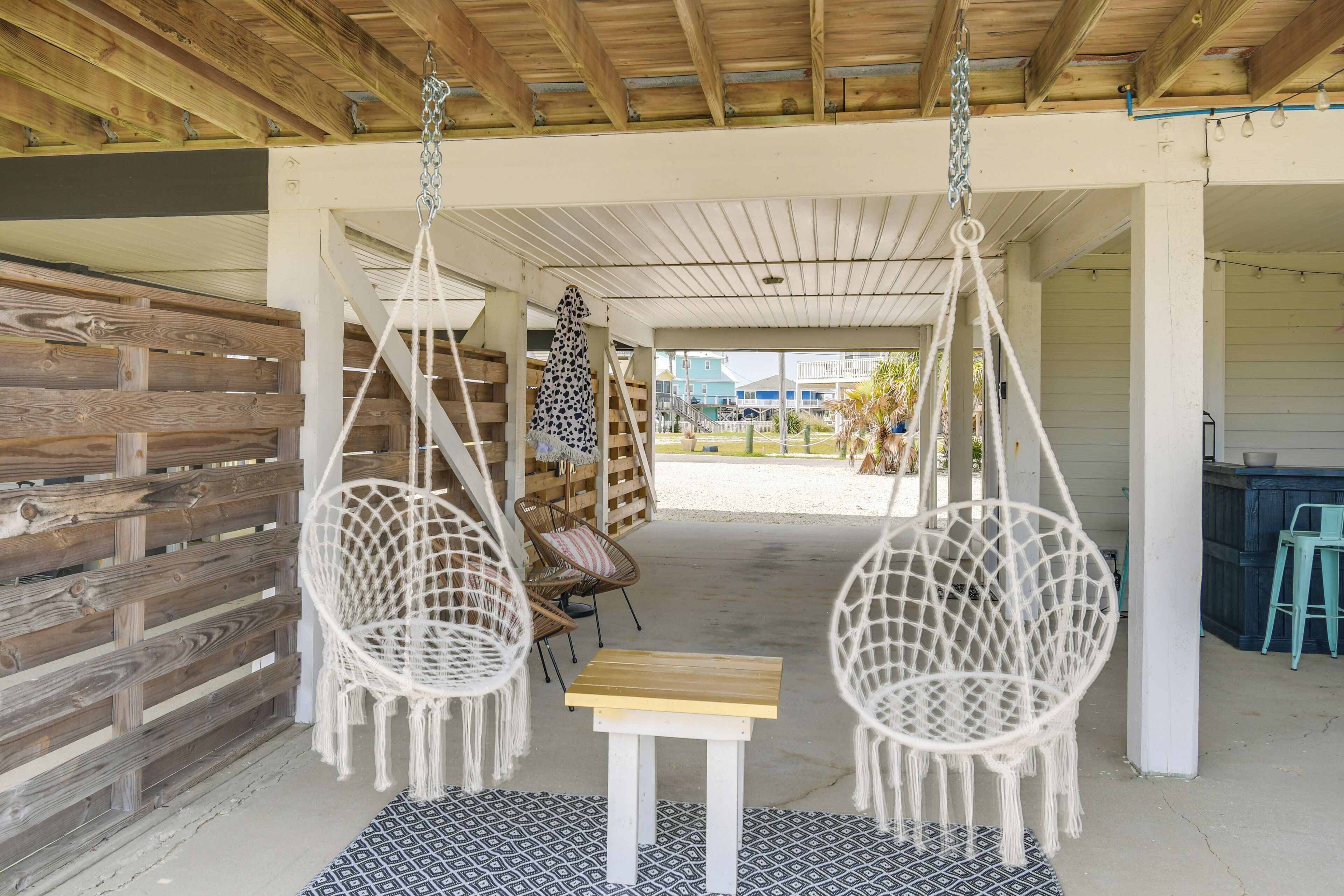 A covered outdoor seating area with macramé chairs, a small table, and a view of the beach and ocean in the background.