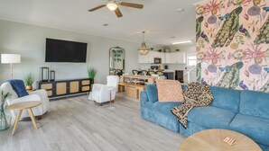 Ein modernes Wohnzimmer mit einem blauen Sofa, einem weißen Sessel und einem Holztisch. Ein Deckenventilator und ein Flachbildfernseher sind an der Wand befestigt.