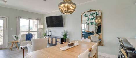Una sala de estar moderna con una mesa de comedor de madera, sillas blancas, un espejo grande con texto motivacional y una araña.