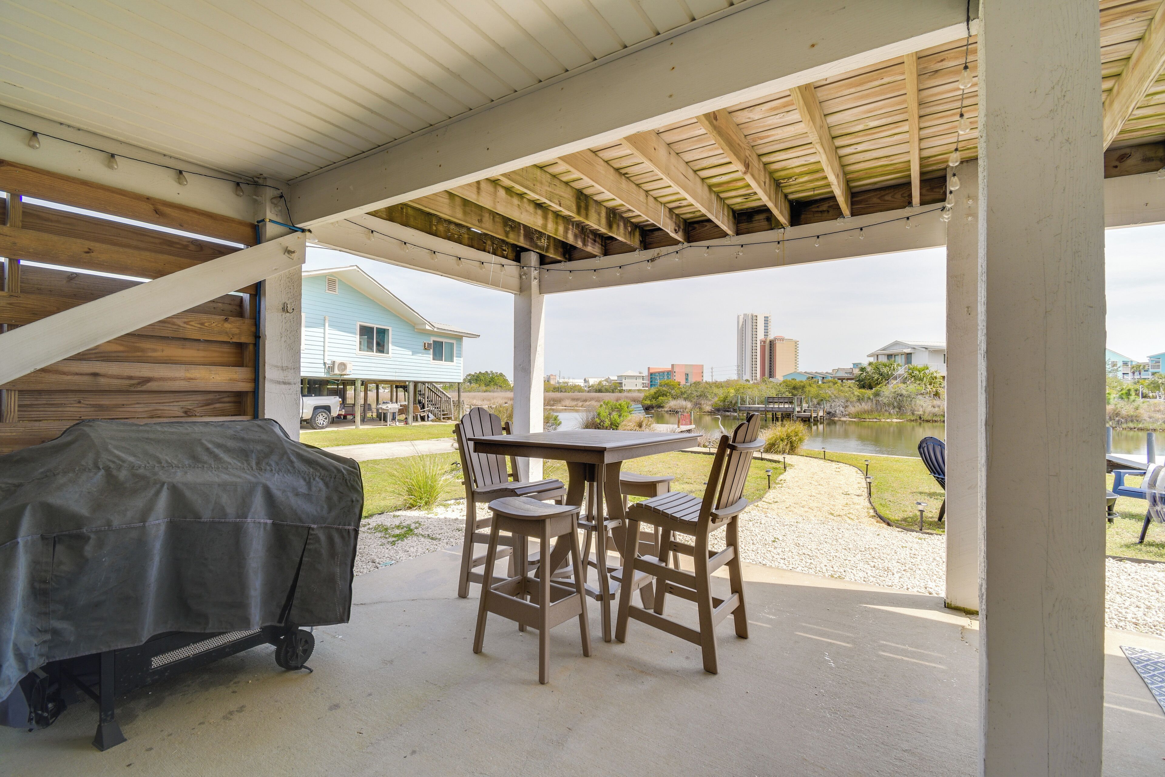 Un patio cubierto con mesa y sillas, una parrilla de barbacoa, y vistas a un lago y edificios al fondo.