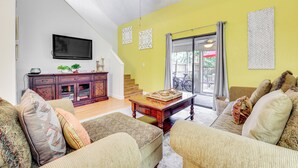 Ein Wohnzimmer mit einer Sofagarnitur, einem Holztisch, einem an der Wand montierten Fernseher und einer Treppe, die zu einem Außenbereich führt.