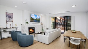 A modern living room with a fireplace, a sofa, blue chairs, a dining table, and a TV mounted on the wall. - New Mtn Modern with Luxurious Amenities | F213 (Avon)