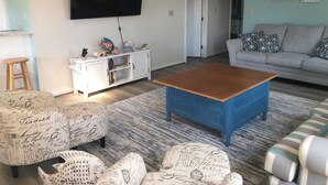 Una sala de estar con televisor de pantalla plana, un aparador blanco con adornos, un sofá gris y otomanas estampadas.