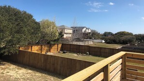 Un patio trasero con terraza de madera, área cercada y una casa al fondo.