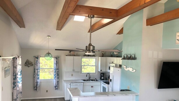 Una cocina con armarios blancos, una isla central con taburetes, un ventilador de techo y una ventana con cortinas.