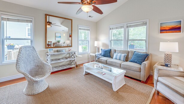 Un salón espacioso con ventilador de techo, una silla de mimbre blanca, una mesa de centro de vidrio, un sofá beige con cojines azules y ventanas grandes.