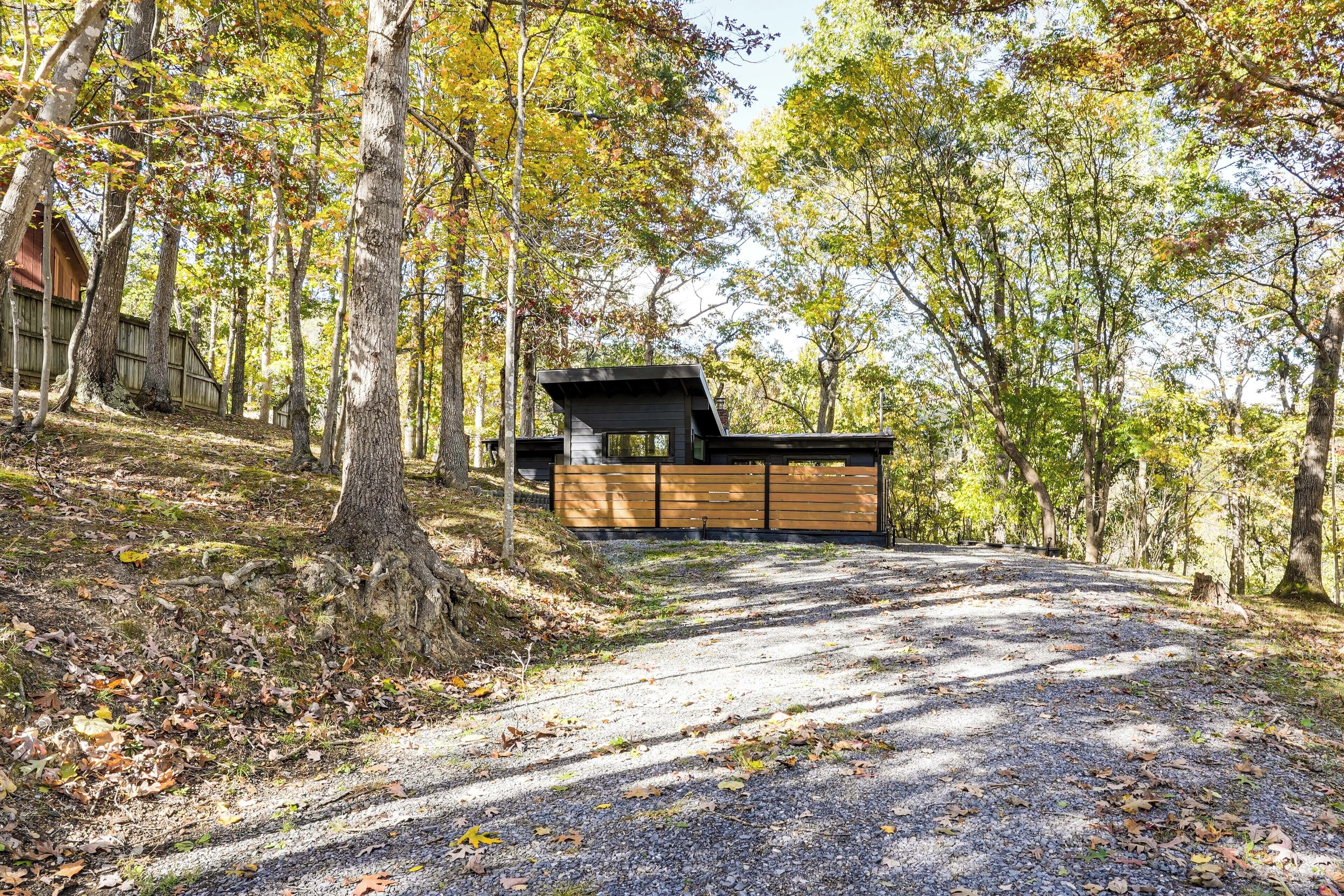 Un chalet rustique avec une allée de gravier menant à une terrasse en bois, entouré d’une forêt aux couleurs automnales.