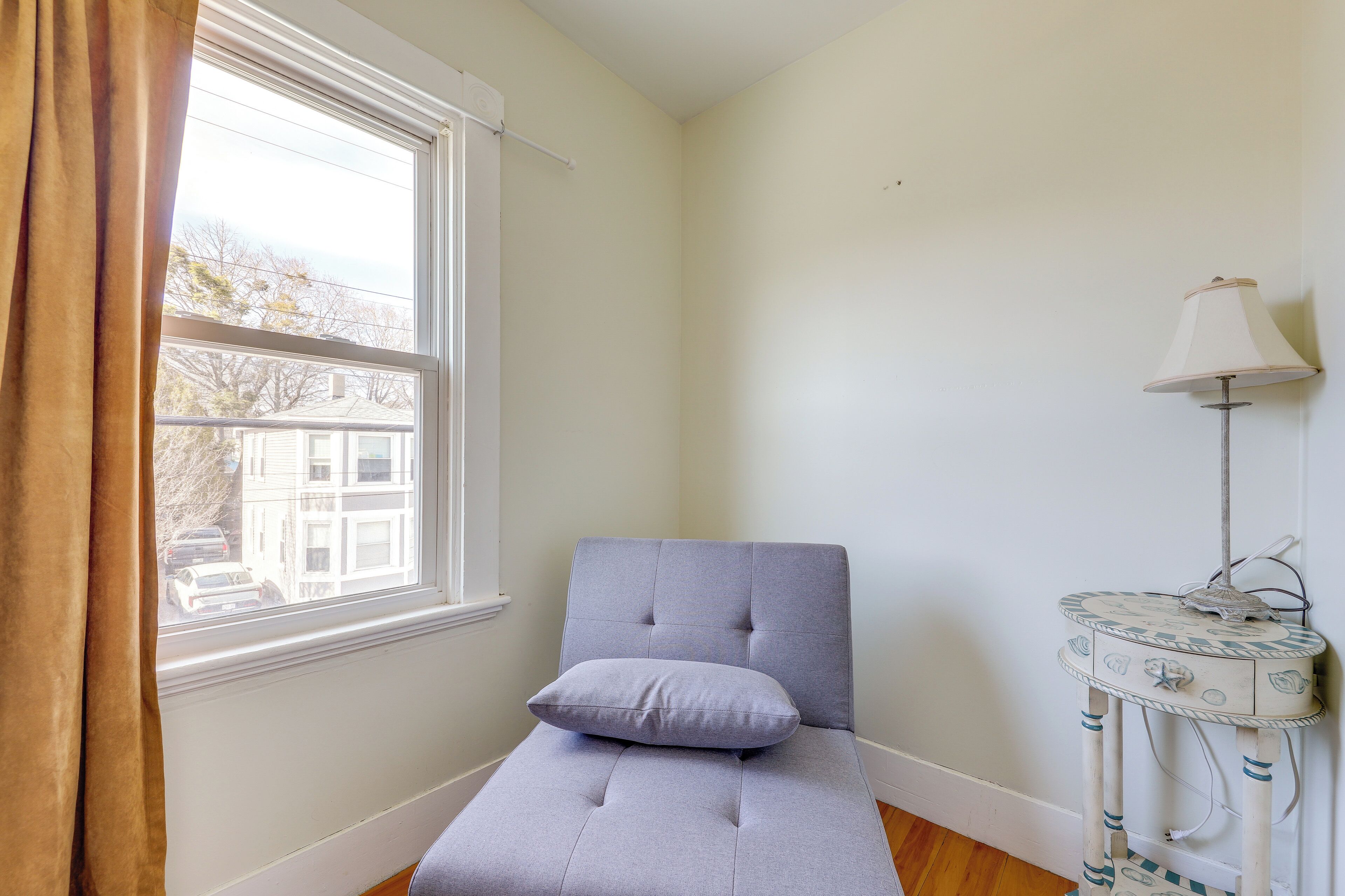 A corner room with a window, a grey cushioned bench, and a small white side table with a lamp.