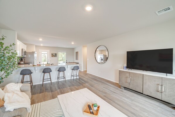Una cocina moderna con una isla central, taburetes de bar, un televisor de pantalla plana grande y una zona de comedor con una mesa y sillas blancas.