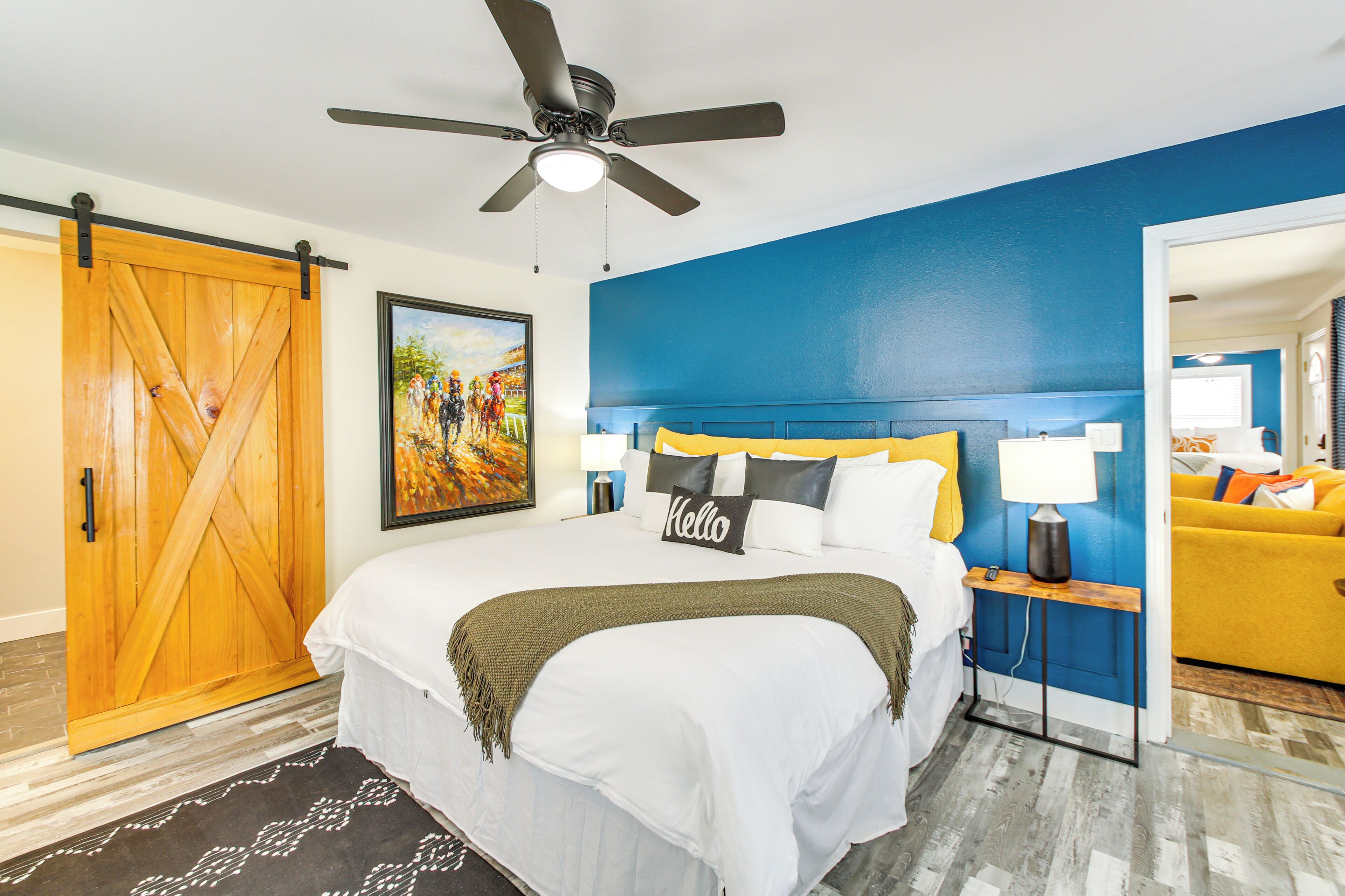 A bedroom with a bed, a ceiling fan, a painting, a nightstand, a lamp, and a yellow sofa.