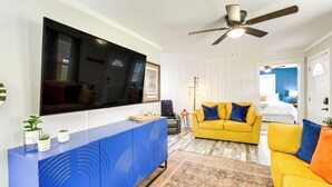 A living room with a flat-screen TV, a blue entertainment unit, a yellow sofa, and a ceiling fan. - Walk to Casino: Home w/ Deck in Hot Springs! (Hot Springs)