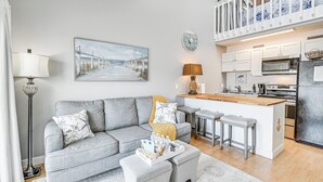 A modern living room with a grey sofa, a coffee table, and a kitchen area with a wooden island and bar stools. - Walk to Beach: Condo w/ Balcony in Murrells Inlet! (Murrells Inlet)