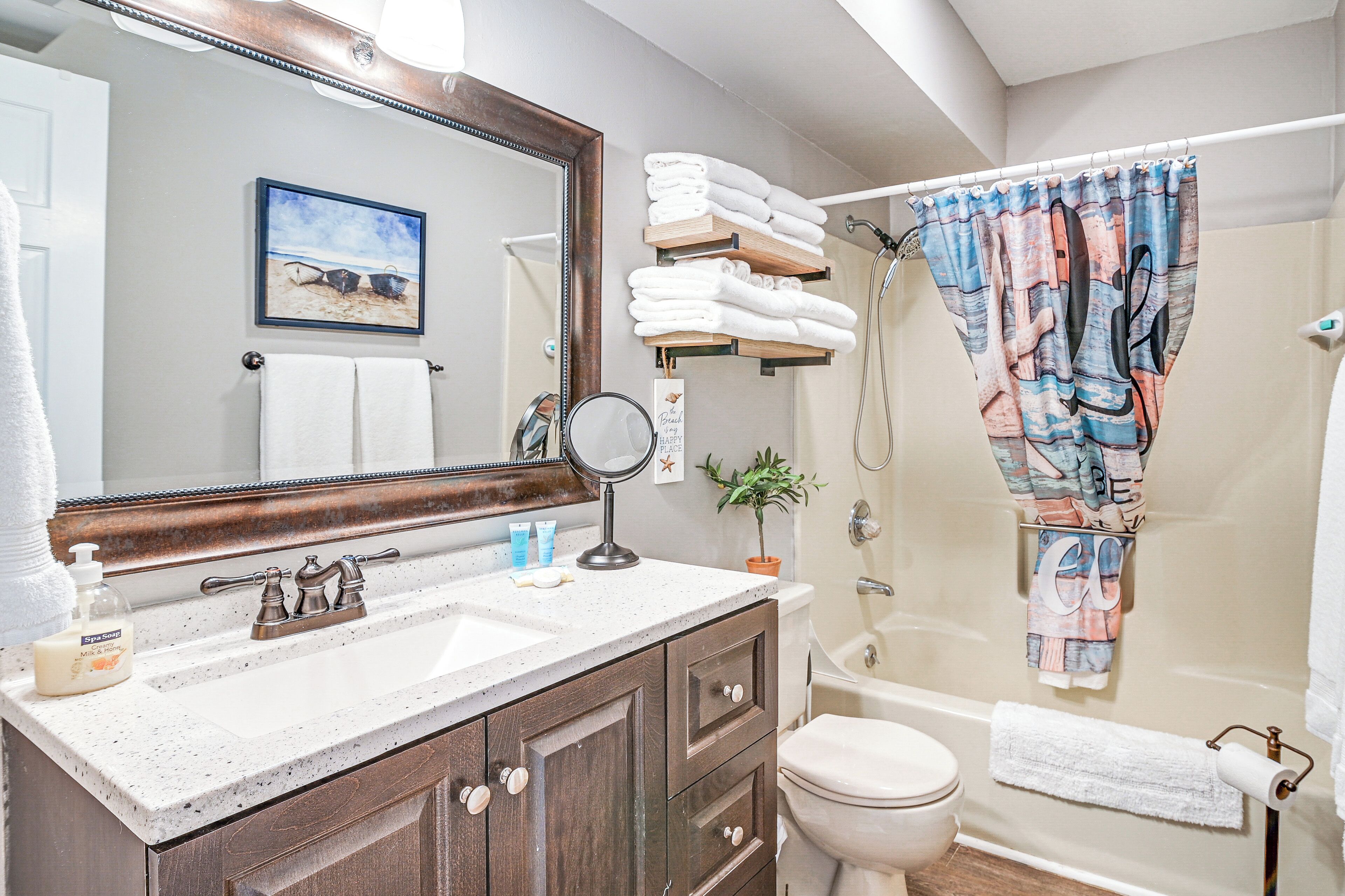 A bathroom with a large mirror, two sinks, a toilet, and a shower. There are towels and toiletries on the counter.