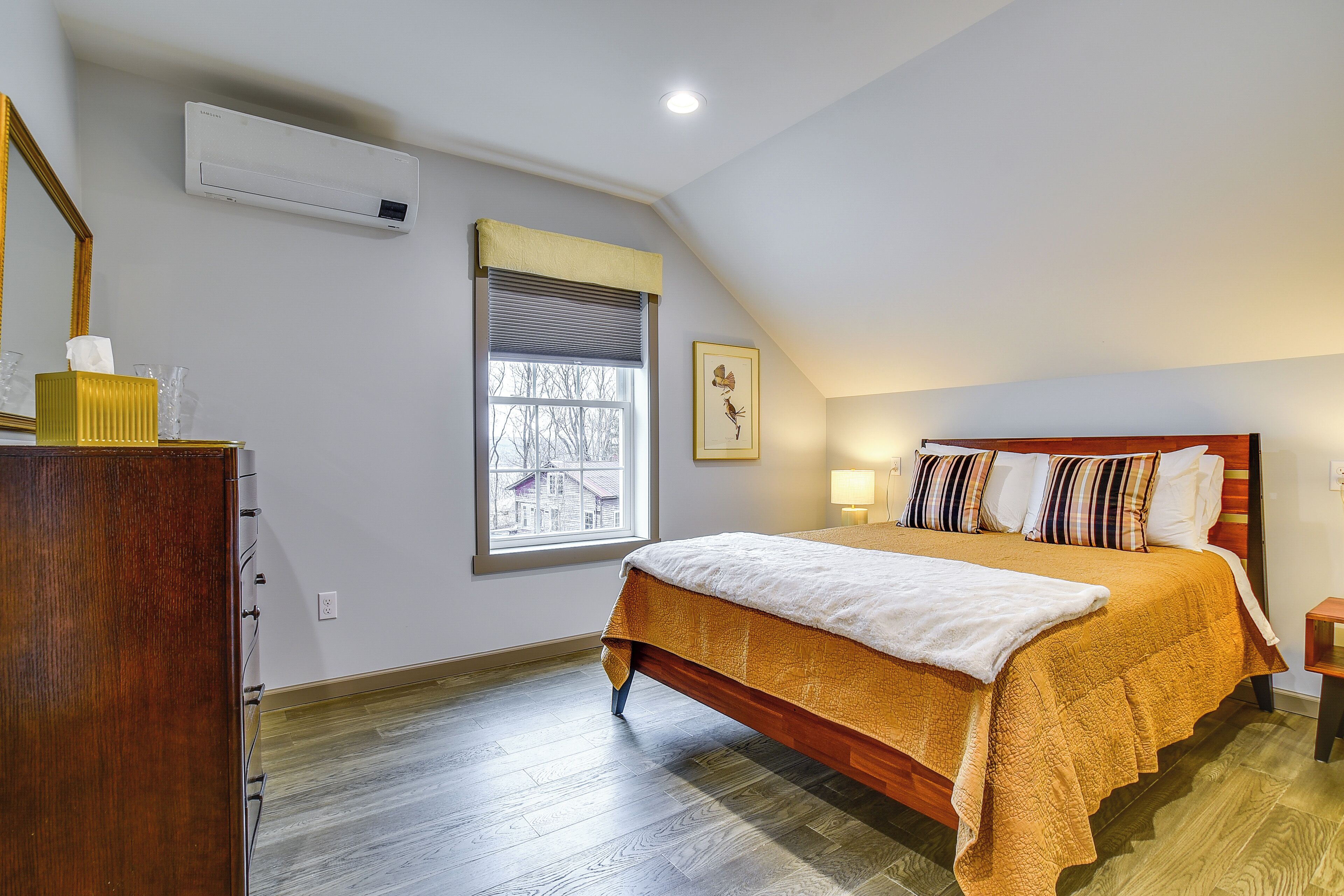 A bedroom with a wooden bed, a window with blinds, an air conditioning unit, and a bedside table with a lamp.