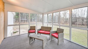 A covered patio with wicker furniture and red cushions, large windows offering a view of a wooded area.