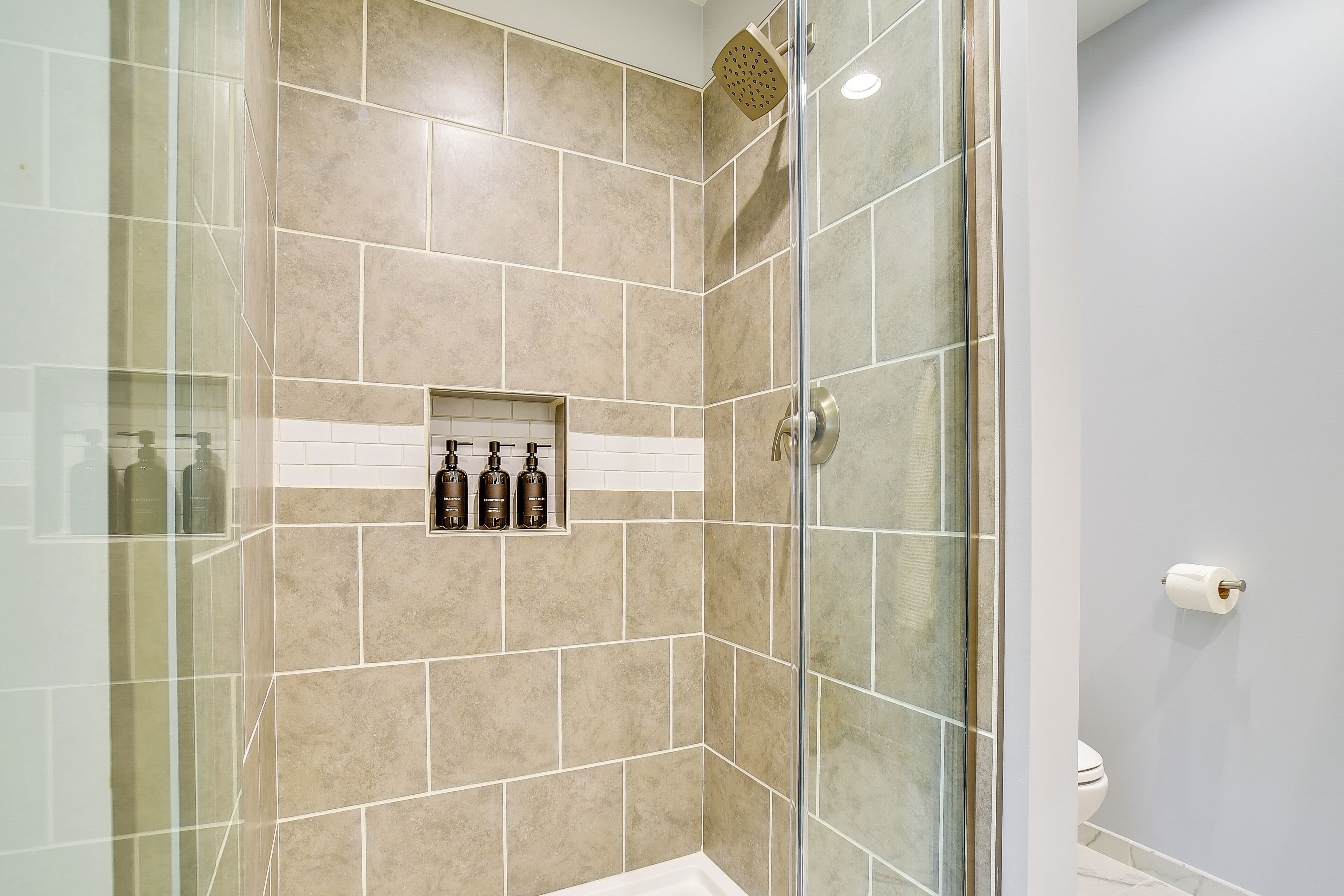 A shower with beige tiles, a glass door, and a shelf with toiletries.