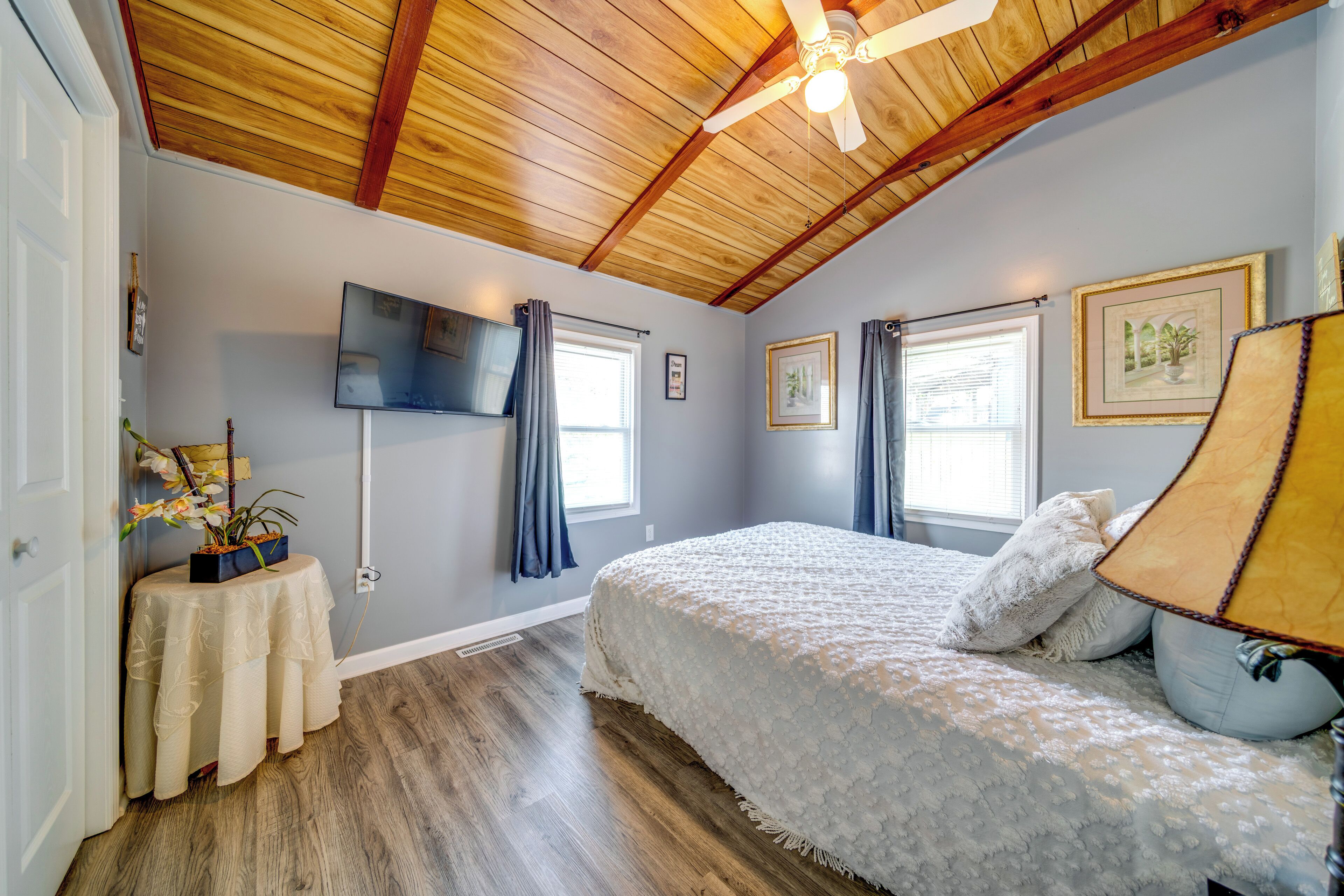 Une chambre avec un plafond en bois, un lit recouvert d’une courtepointe blanche, un lampadaire et un téléviseur à écran plat fixé au mur.