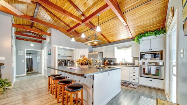 Une cuisine moderne avec des meubles blancs, un grand îlot central doté de tabourets, des poutres en bois au plafond et une plante suspendue.