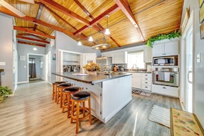 A modern kitchen with white cabinets, a large island with bar stools, wooden ceiling beams, and a hanging plant.