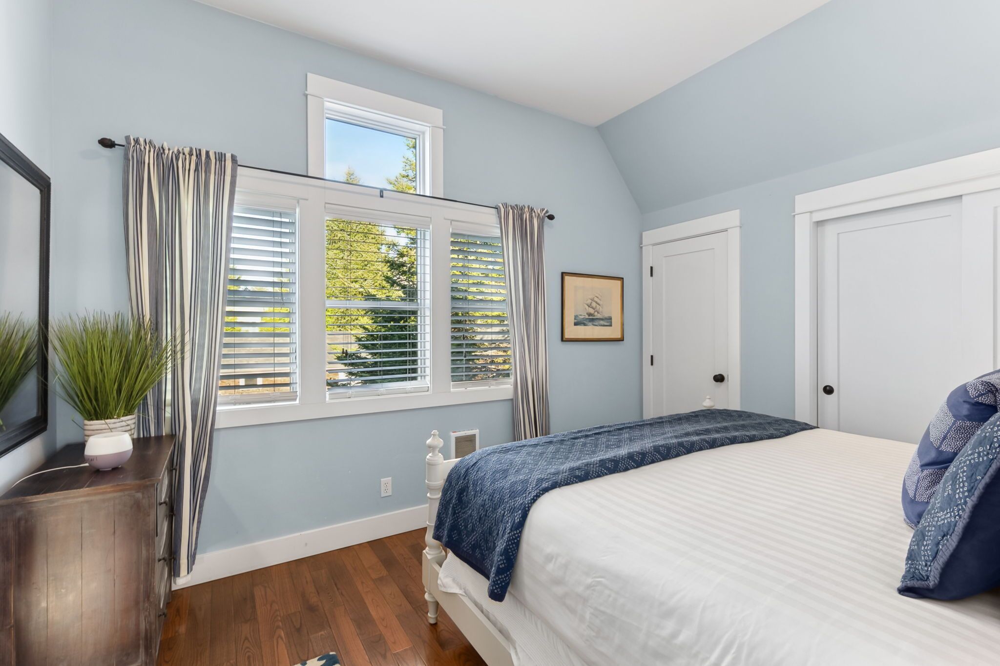 A bedroom with a bed, a window with curtains, a door, and a small plant on a dresser.
