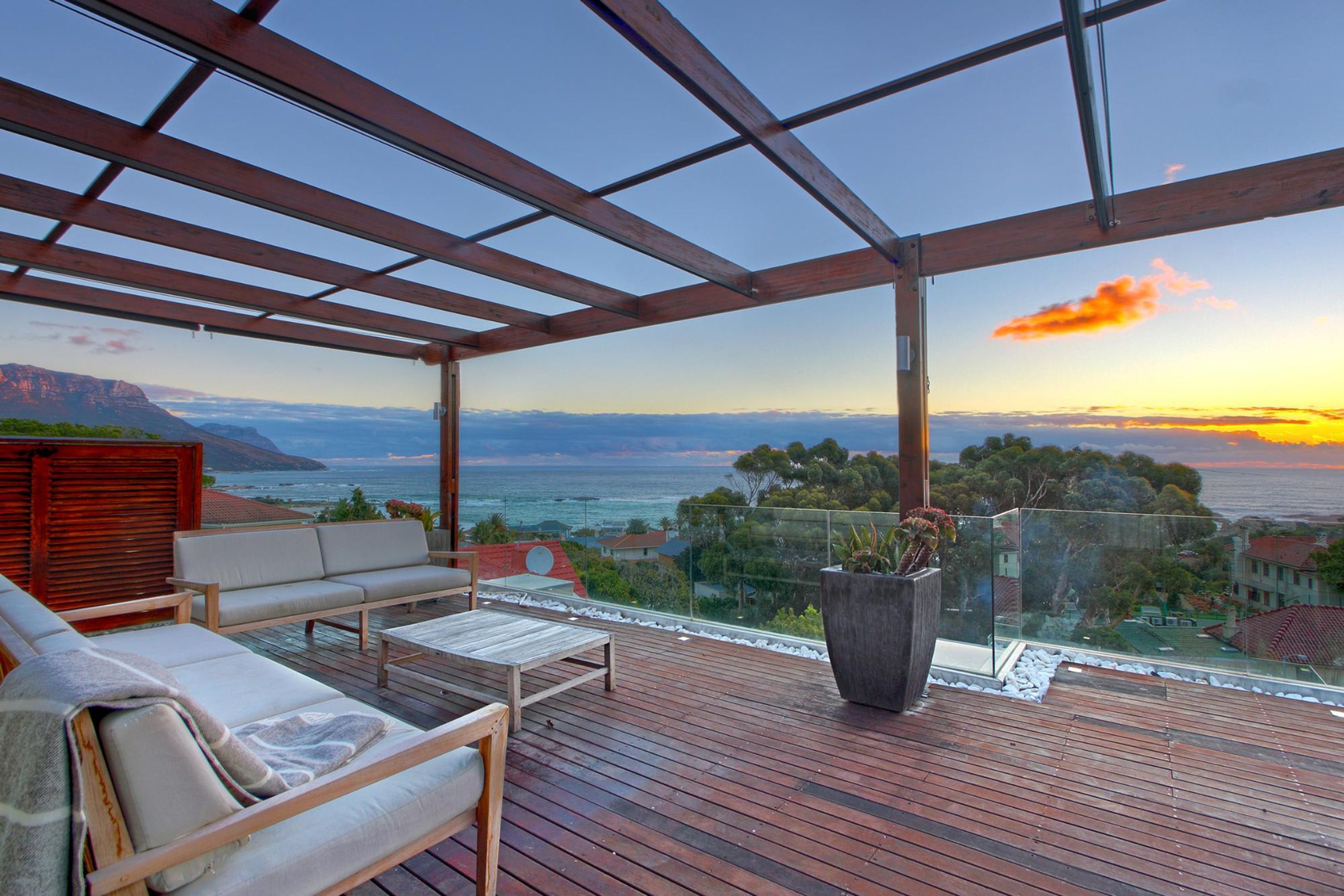 Eine Holzterrasse mit Sitzgelegenheiten im Freien, ein großer Kübel mit Pflanze und Blick auf das Meer und die Berge bei Sonnenuntergang.