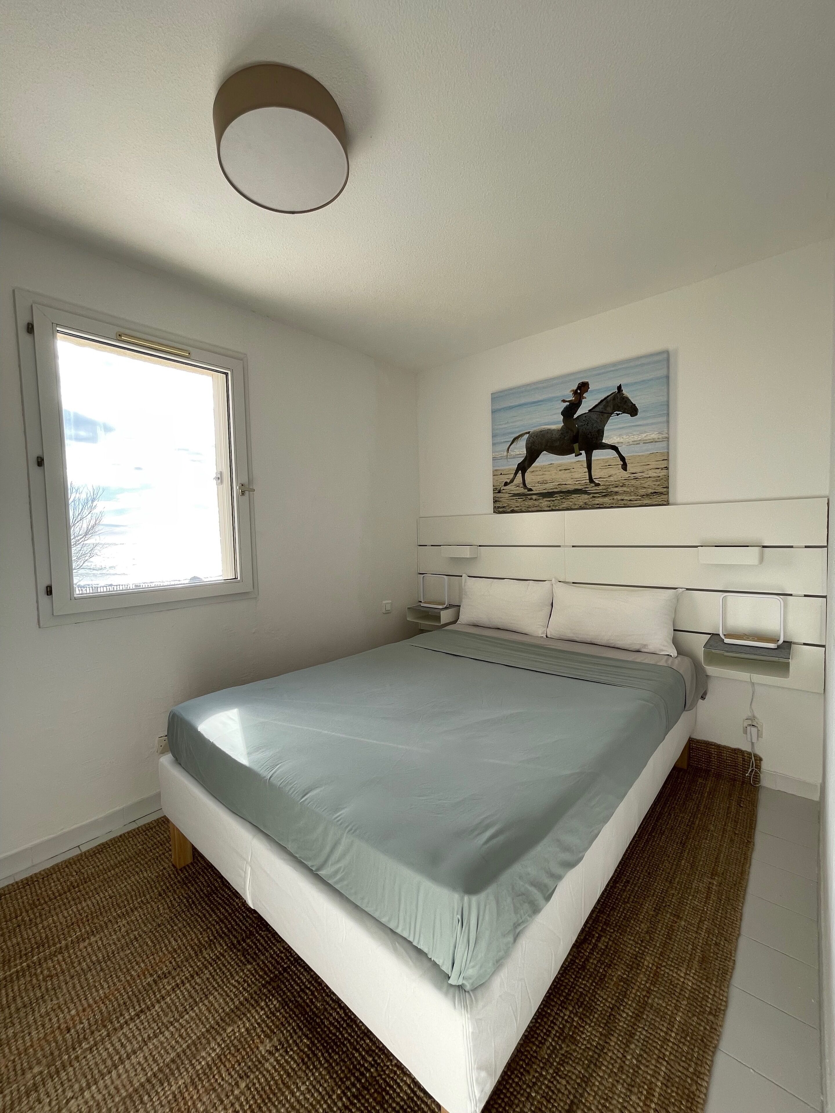 Ein Schlafzimmer mit Bett, Fenster, einem Bild und einem Nachttisch.