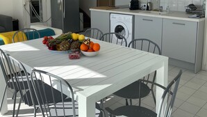 A modern kitchen with white cabinets, a stainless steel refrigerator, a washing machine, and a dining area with a white table and metal chairs. - Maison Front de Mer Aresquiers | Direct beach access | 4 bedrooms (Frontignan)