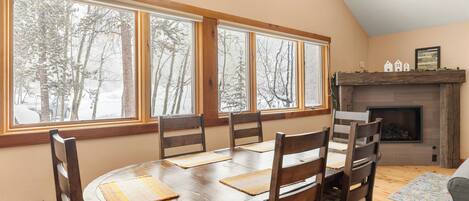 A dining area with a round table, wooden chairs, and large windows with a view of trees.
