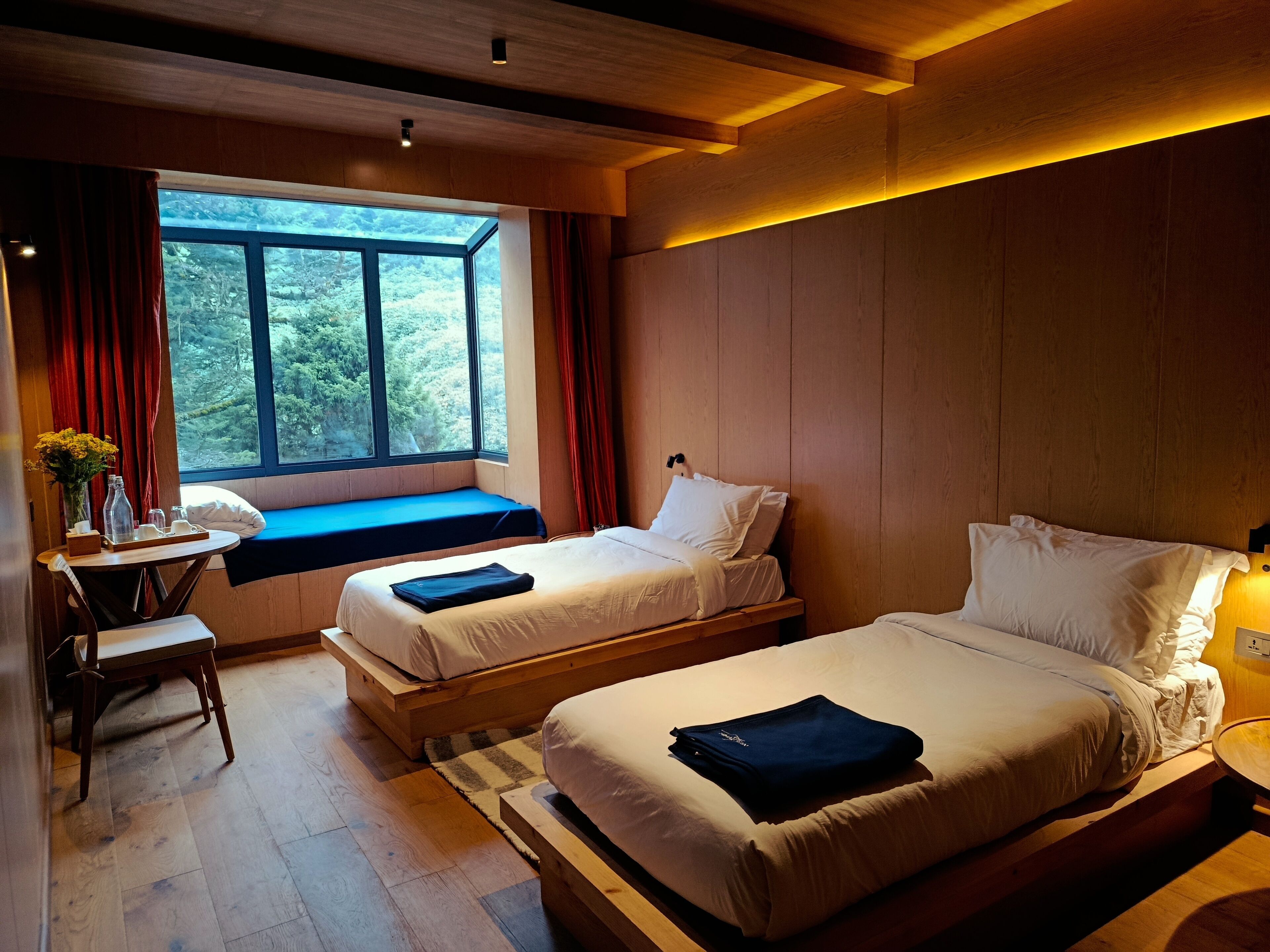 A hotel room with two beds, a desk with a chair, a window with curtains, and a view of trees.