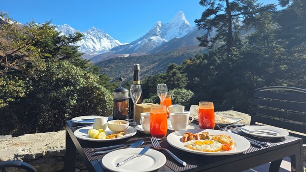 A breakfast table set outdoors with a mountain view, including a bottle, glasses, and plates of food. -  The Himalayan Tyangboche (Lukla)