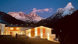 A modern building with large windows and a stone facade, situated in a mountainous area with snow-capped peaks in the background. -  The Himalayan Tyangboche (Lukla)