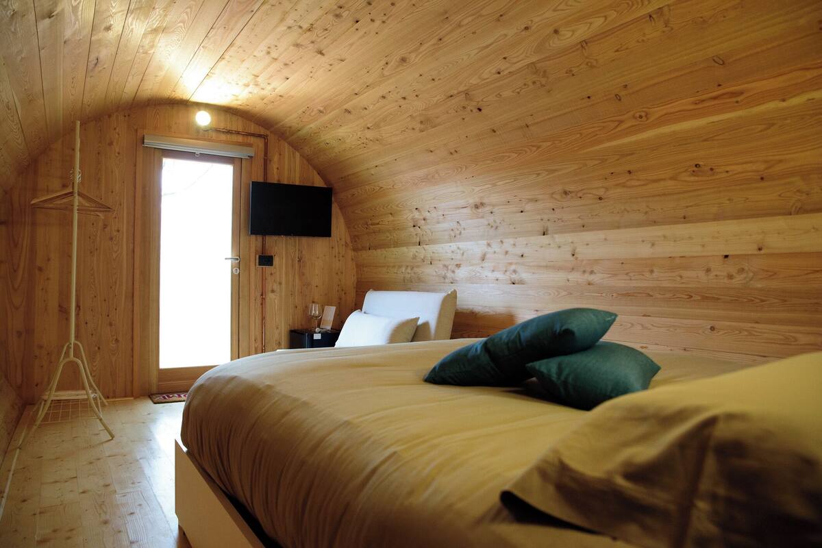 A cozy, wooden-paneled bedroom with a bed, pillows, a flat-screen TV, and a door leading to a bright room.