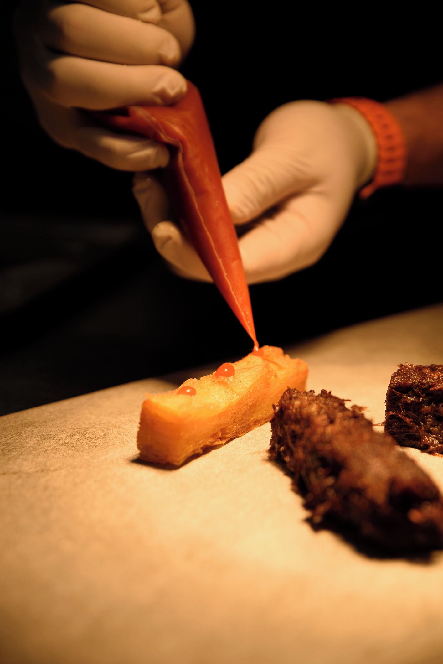A person using a red piping bag to apply a substance to a food item on a cutting board.