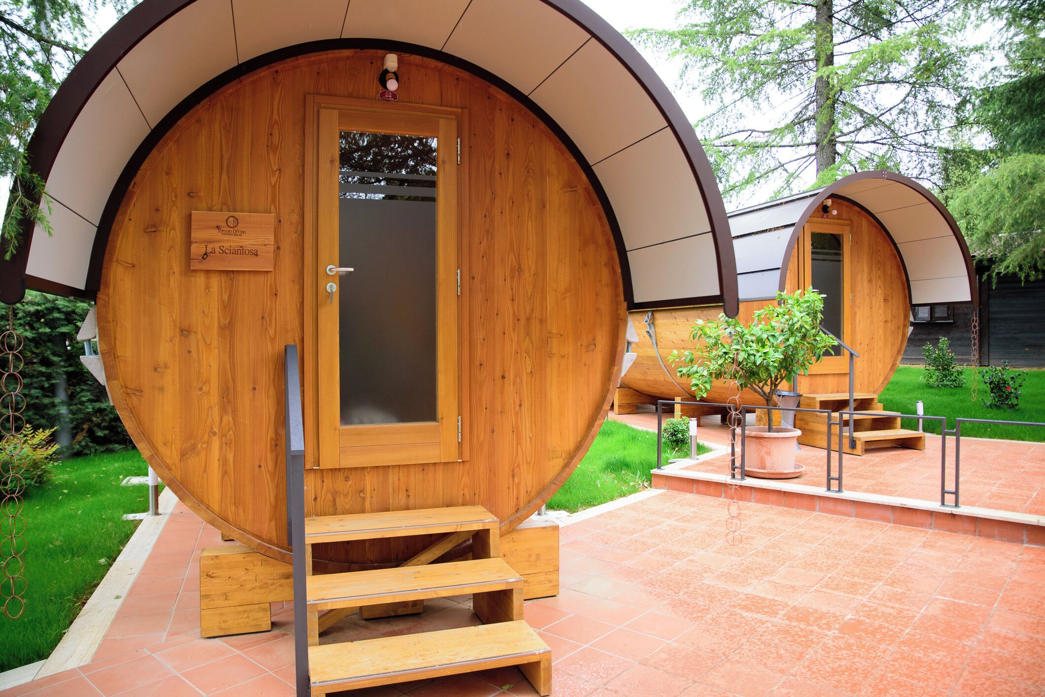 Two wooden barrel-shaped cabins with wooden steps leading to the entrance, surrounded by a garden and a paved area.