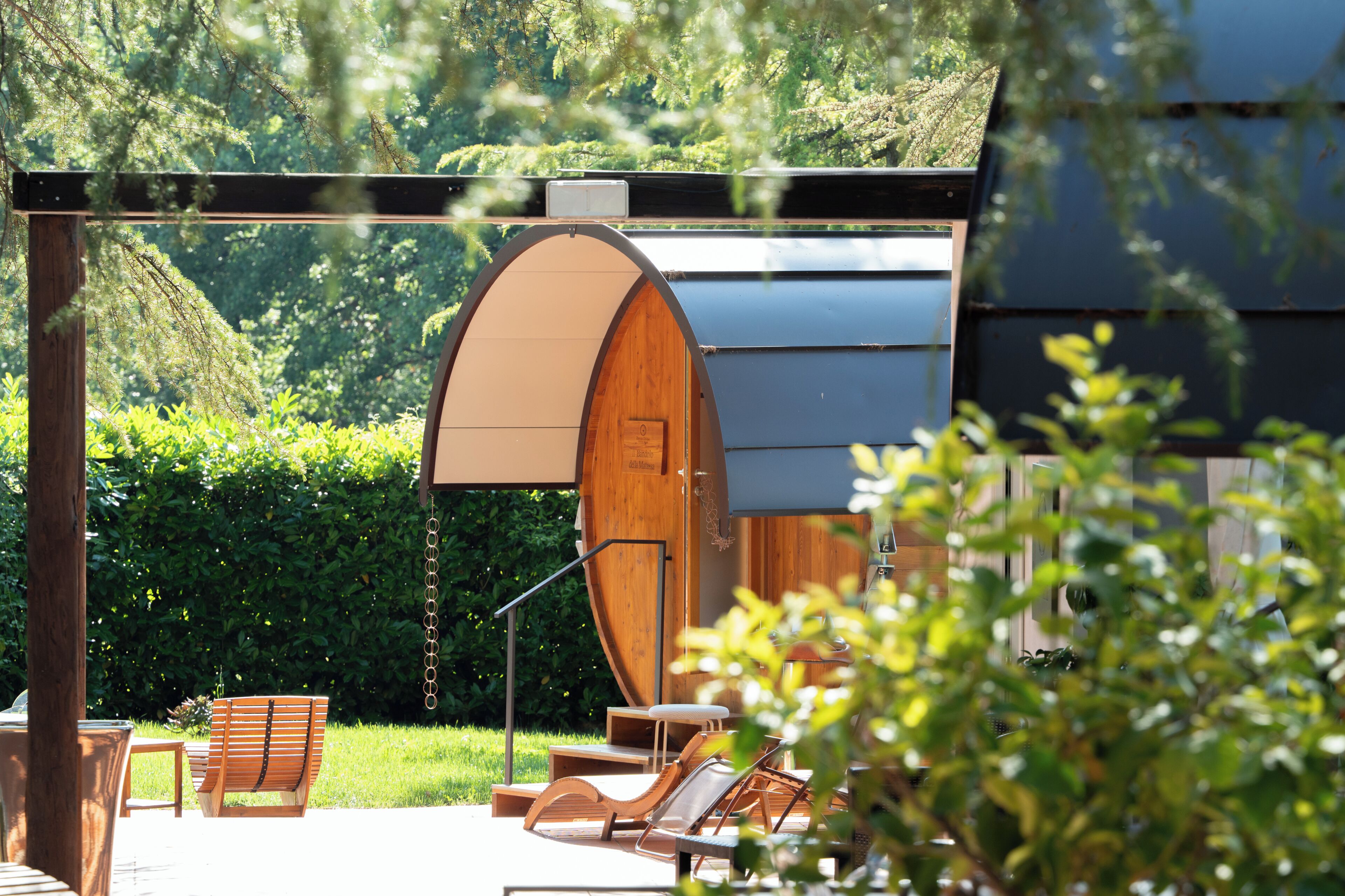 A wooden barrel-shaped structure, outdoor seating with chairs and a table, and a hedge in the background.