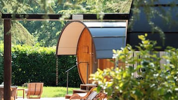 A wooden barrel-shaped structure, outdoor seating with chairs and a table, and a hedge in the background.