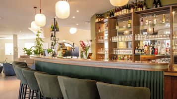Eine Bar mit Theke, Barhocker, Regalen für Flaschen und Gläser sowie hängenden Pendelleuchten.