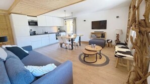 Ett modernt vardagsrum med en grå soffa, en matplats med bord och stolar, ett kök med vita skåp och en stor träskulptur föreställande ett träd.