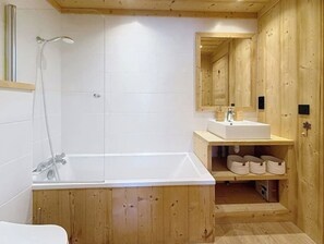 Ett badrum i en stuga med träpanel, ett vitt badkar, en dusch och ett handfat med en spegel ovanför.