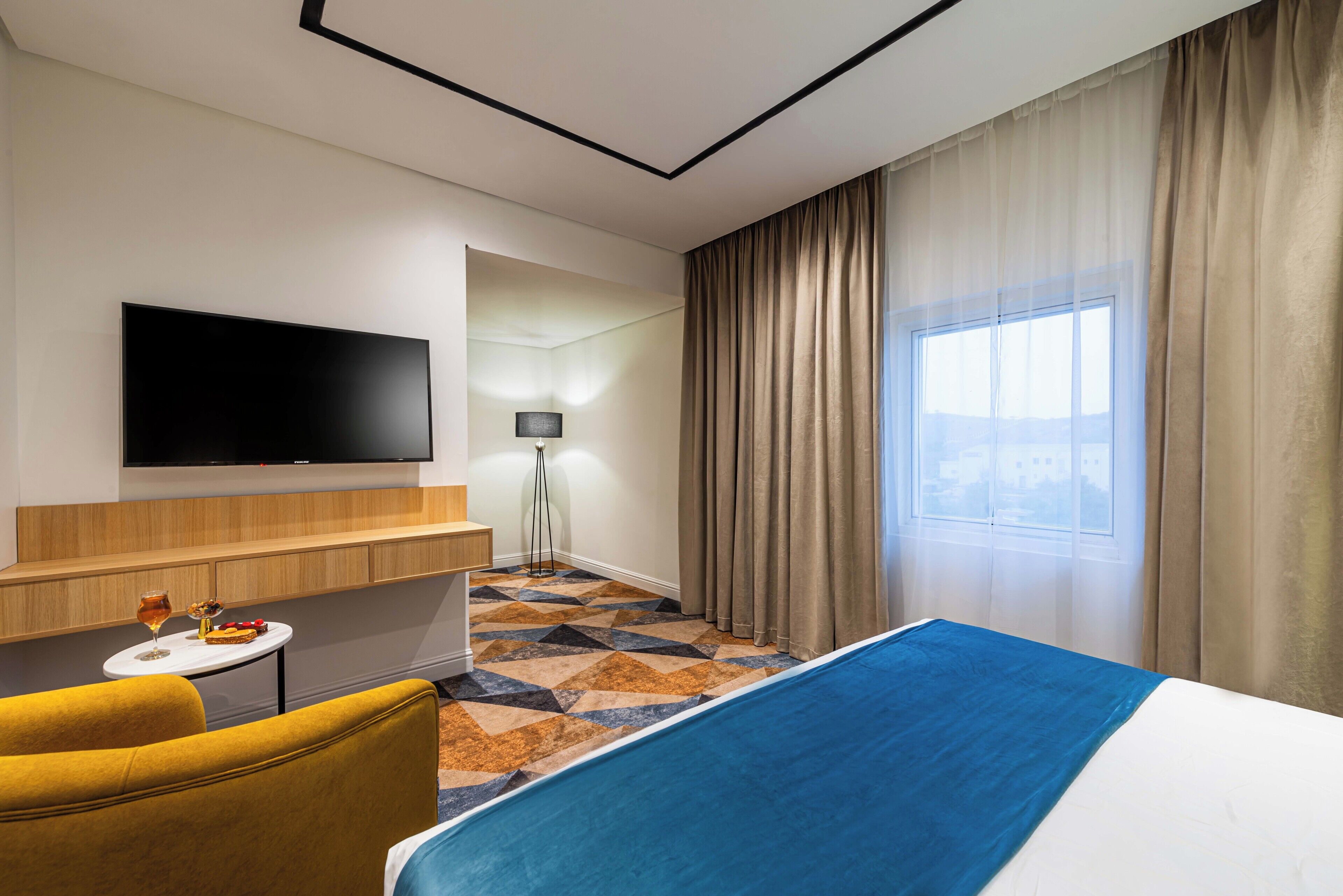Habitación de hotel con cama, televisor montado en la pared, una pequeña mesa con una copa de vino, un sillón amarillo y alfombra estampada.