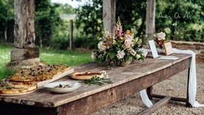 Una mesa de madera rústica con un arreglo floral y varios platos, ubicada al aire libre con árboles de fondo.