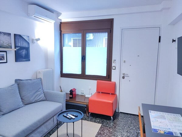 A compact living space with a grey sofa, a red chair, a small table with remotes, a window with blinds, and a door. - LIV - Cozy Nomad Retreat (Athens)