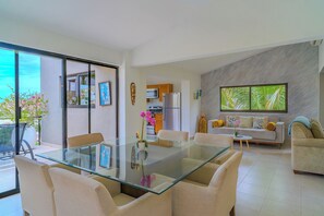 A modern living space with a glass dining table, beige sofas, and a kitchen area visible through an open door.