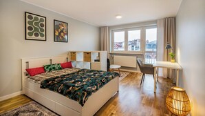 Une chambre moderne avec un lit, des tables de chevet, un bureau et une vue sur la ville grâce à de grandes fenêtres.