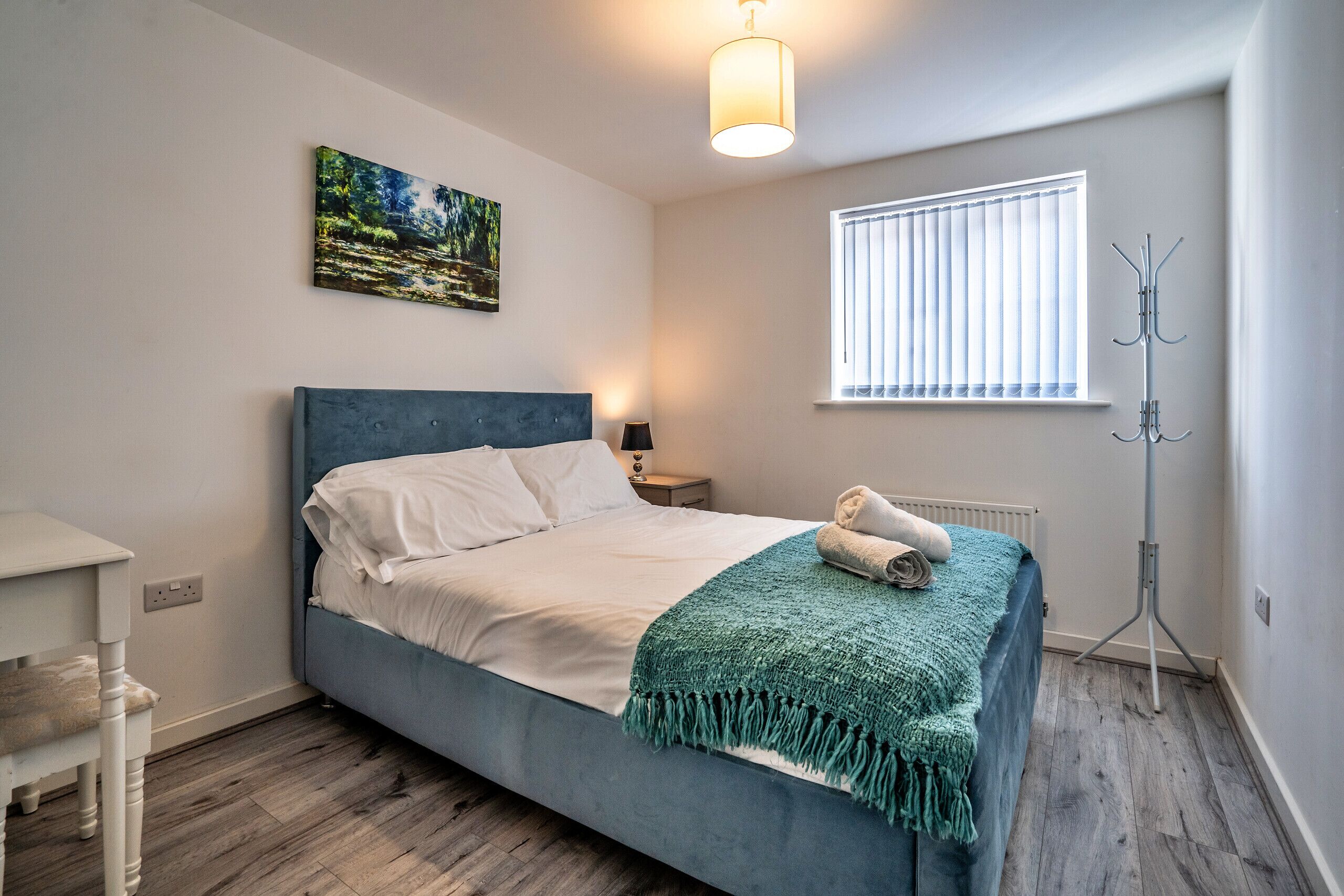 A bedroom with a bed, a bedside table, a lamp, a window with blinds, a coat rack, and a painting on the wall.
