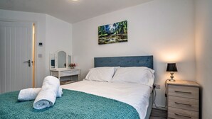 A neatly made bed with a teal blanket and rolled towels, a bedside table with a lamp, a wall-mounted mirror, and a painting of a landscape. - Aldridge Square (Birmingham)